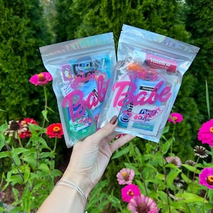 May include: Two clear plastic bags filled with various beauty products, held in front of a garden with pink flowers. Each bag has a pink "Babe" label. One bag contains a green bottle of soap. The other bag contains a tube of Vaseline.
