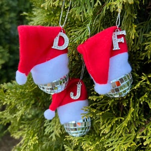 May include: Three red and white Santa hat ornaments with silver glitter disco balls and silver initial charms. The initials are D, F, and J.