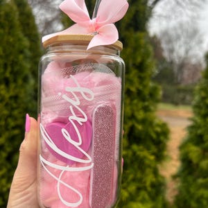 May include: A clear glass jar with a bamboo lid and a pink ribbon. The jar contains pink cotton balls, a pink heart-shaped sponge, and a pink nail file. The jar is labeled "Alexis".
