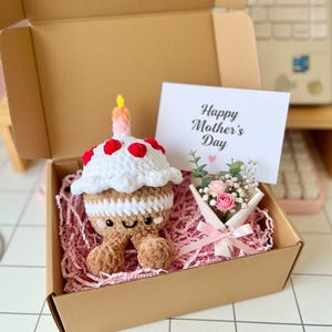 May include: A crocheted cupcake-shaped plush toy with a candle, red accents, and a smiling face, presented in a gift box with a "Happy Mother's Day" card and a small bouquet of flowers. The box is filled with pink shredded paper.