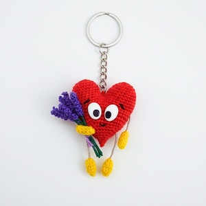 Pode incluir: Um chaveiro vermelho em forma de coração de crochê, com olhos grandes e um rosto sorridente. O coração segura um buquê de flores roxas e tem pés amarelos. O chaveiro é preso a um anel de metal prateado.