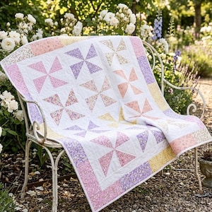 May include: A handmade quilt featuring a white background with repeating pinwheel blocks in pastel floral patterns of pink, purple, and yellow. The quilt has a matching floral border and is draped over a white metal garden bench in a flower garden.