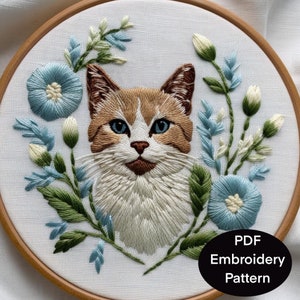Peut inclure: Broderie d'un portrait de chat dans un cercle en bois. Le chat a un pelage brun et blanc avec des yeux bleus, entouré de fleurs bleues et de feuilles vertes. Le texte "PDF Embroidery Pattern" est dans un ovale noir.