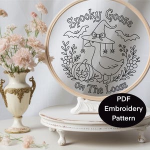 Pu&ograve; includere: Un motivo per ricamo in bianco e nero in un telaio di legno. Il disegno presenta un'oca con un cappello da strega, una zucca di Halloween, pipistrelli e la scritta "Spooky Goose On The Loose". Un ovale nero con la scritta "PDF Embroidery Pattern" si trova in basso a destra.
