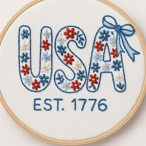 K&ouml;nnte beinhalten: Gesticktes Wandbild mit dem Wort "USA" in Blau, gef&uuml;llt mit roten, wei&szlig;en und blauen Blumenmustern. Der Text "EST. 1776" ist darunter gestickt. Das Design befindet sich in einem Holzreifen.