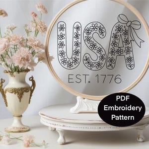 K&ouml;nnte beinhalten: Ein runder Stickrahmen mit dem Wort "USA" in Schwarz, verziert mit floralen Mustern. Der Text "EST. 1776" steht unter den Buchstaben. Ein schwarzes Oval mit dem Text "PDF Embroidery Pattern" befindet sich unten. Links ist eine Blumenvase.