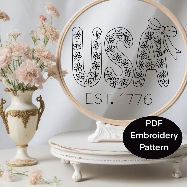 K&ouml;nnte beinhalten: Ein runder Stickrahmen mit dem Wort "USA" in Schwarz, verziert mit floralen Mustern. Der Text "EST. 1776" steht unter den Buchstaben. Ein schwarzes Oval mit dem Text "PDF Embroidery Pattern" befindet sich unten. Links ist eine Blumenvase.