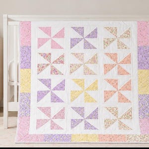 May include: A rectangular baby quilt featuring a pinwheel pattern made of various floral and solid pastel fabric squares in shades of pink, yellow, and lavender. The quilt is bordered by a white frame and a perimeter of alternating colored squares.