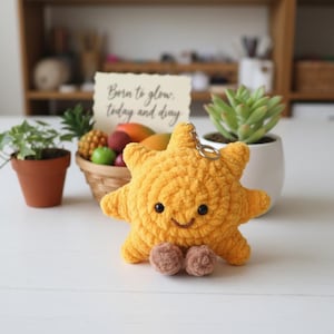 May include: A crocheted yellow sun-shaped keychain with a smiling face and brown feet. The keychain has a silver ring attached. The text "Born to glow, today and day" is on a card in the background.
