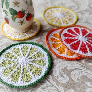 Könnte beinhalten: Eine Sammlung handgefertigter Häkeluntersetzer in Form von Zitrusfruchtscheiben. Die Untersetzer umfassen eine Limette, Zitrone, Orange und Grapefruit, jeweils mit detaillierten Nähten und leuchtenden Farben. Eine Teetasse mit floralem Design ruht auf einem passenden Untersetzer.
