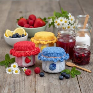 Op de afbeelding: Drie kleine potjes met kleurrijke gehaakte deksels, rood, geel en blauw, staan op een houten oppervlak. Elk potje heeft een fruitontwerp op de voorkant. Schalen met verse bessen, citroenen en madeliefjes omringen de potjes, wat op zelfgemaakte jam duidt.