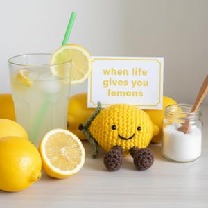 May include: A glass of lemonade with a green straw and lemon slice, next to fresh lemons and a crocheted lemon character with the text "when life gives you lemons". A jar of sugar and a wooden spoon are also present.