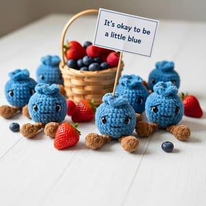 May include: A whimsical display features several blue crochet blueberry characters with smiling faces and brown feet, alongside fresh strawberries and blueberries. A small basket holds more berries, and a sign reads "It's okay to be a little blue."
