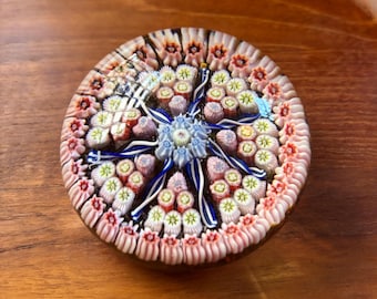May include: A round, glass paperweight with a floral design. The center features a blue and white flower, surrounded by pink, red, and yellow floral patterns. The edges are lined with a pink and white pattern. The paperweight is set on a wooden surface.