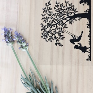 May include: A wooden board with a black silhouette of a tree with two children on a swing. There is a sprig of lavender in the bottom left corner of the image.