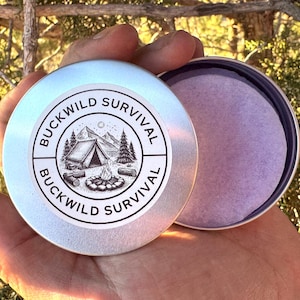 May include: A silver tin with the words "BUCKWILD SURVIVAL" encircling a graphic of a tent, trees, and a campfire. The tin is open, revealing a purple pad inside. The tin is held in a hand.