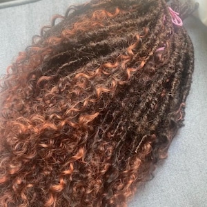 May include: A bundle of dark brown and auburn curly hair extensions. The hair is styled in individual locs with a natural, textured appearance. Pink elastics secure the ends of some locs, adding a pop of color.