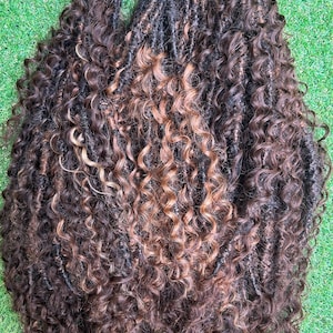 May include: A bundle of synthetic hair extensions featuring dark brown and copper-toned highlights. The hair is styled in long, textured locs with loose, spiral curls at the ends. Each section is secured at the top with a small pink string tie.