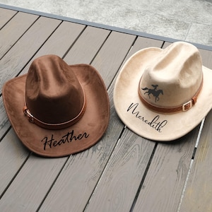 May include: Two cowboy hats, one brown and one beige, rest on a wooden surface. The brown hat has the name "Heather" in black script. The beige hat features a silhouette of a rider on a horse and the name "Meredith". Both hats have brown leather bands.