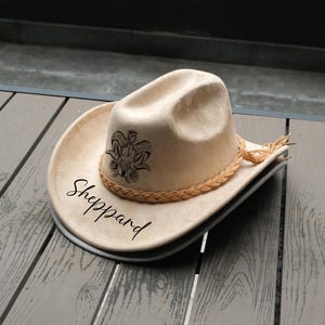 Puede incluir: Un sombrero de vaquero beige con una banda trenzada y un diseño decorativo de calavera de carnero y rosas. La palabra "Sheppard" está escrita en una fuente cursiva en el borde. El sombrero tiene un ala ancha y una forma clásica.