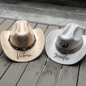 May include: Two cowboy hats. One is tan with a horseshoe and landscape design, the name "Victoria" is printed below. The other is light gray with a Texas outline design, and the name "Jennifer" is printed below.