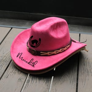 May include: A vibrant pink cowboy hat with a brown leather band and decorative accents. The hat features a black horseshoe and horse head design, with the name "Meredith" in script. A stylish accessory.