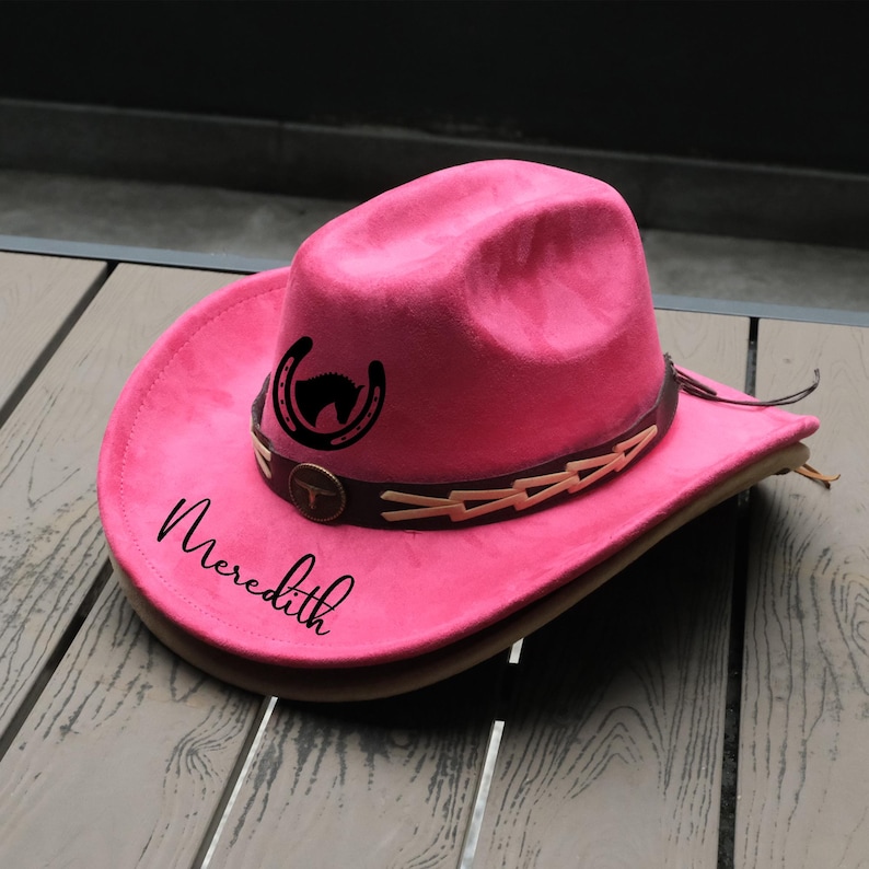 May include: A vibrant pink cowboy hat with a brown leather band and decorative accents. The hat features a black horseshoe and horse head design, with the name "Meredith" in script. A stylish accessory.