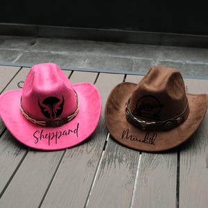 May include: Two cowboy hats. One is bright pink with a black graphic and the name "Sheppard" in black script. The other is brown with a graphic and the name "Meredith" in black script. Both have a decorative band.