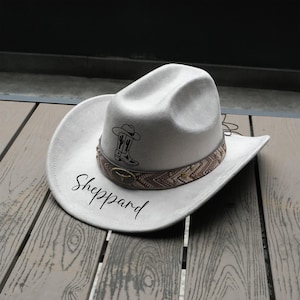 May include: A light gray cowboy hat with a decorative band featuring a brown and tan pattern. The hat has a black graphic of a cowboy boot and cactus. The name "Sheppard" is written in a script font on the hat.