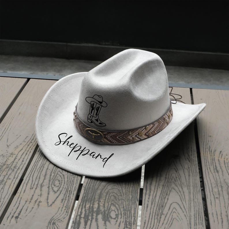 May include: A light gray cowboy hat with a decorative band featuring a brown and tan pattern. The hat has a black graphic of a cowboy boot and cactus. The name "Sheppard" is written in a script font on the hat.