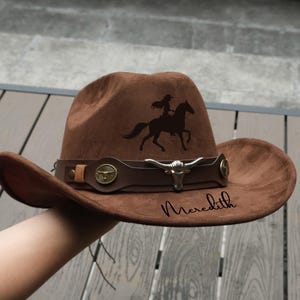 Könnte beinhalten: Brauner Cowboyhut mit einer Silhouette eines Reiters auf einem Pferd. Der Hut hat ein Lederband mit einem silbernen Bullenkopf und dekorativen Akzenten. Der Name "Meredith" ist auf dem Hut geschrieben.