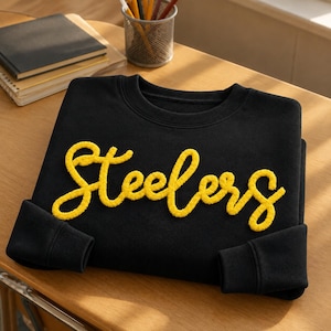 May include: A black long-sleeve crewneck sweatshirt featuring the word Steelers written in a yellow, textured, raised chenille-style script font across the chest. The garment has a ribbed collar and ribbed cuffs on the long sleeves.