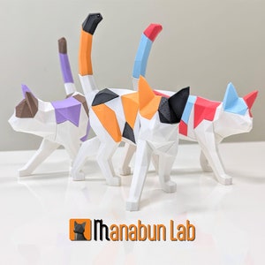 Puede incluir: Tres figuritas de gato de diseño geométrico hechas de plástico blanco con diferentes acentos de color. Los gatos están en fila, mirando hacia la derecha, con el gato de atrás teniendo un esquema de color negro, naranja y blanco, el gato del medio teniendo un esquema de color morado, marrón y blanco, y el gato de adelante teniendo un esquema de color rojo, azul y blanco. Los gatos están sobre una superficie blanca con el texto "Thanabun Lab" en letras naranjas sobre un fondo blanco con una silueta de gato negro en la esquina.