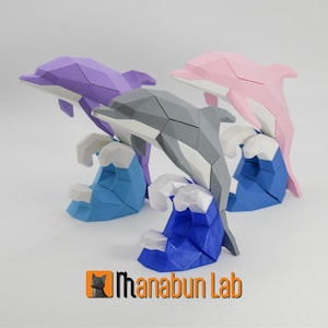 May include: Three origami-style dolphin sculptures in purple, pink, and gray, each leaping over a blue and white wave. The sculptures are displayed on a white surface. The logo "Ihanabun Lab" is in the bottom right corner.