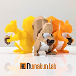 May include: Three 3D printed squirrel figurines holding acorns. The squirrels are made of brown, orange, and yellow plastic. The figurines are standing on a white surface.