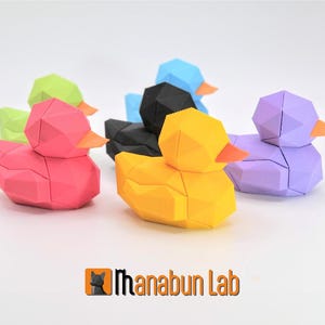 May include: A collection of geometric origami-style duck figurines in various colors, including pink, yellow, black, blue, green, and purple. Each duck has an orange beak. The logo "Manabun Lab" is visible at the bottom.
