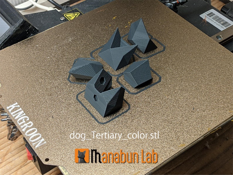 May include: A 3D printer bed with several dark gray geometric shapes. The shapes vary in design, including angular and faceted forms. The text "dog_Tertiary_color.stl" and the logo for "Ihanabun Lab" are visible.