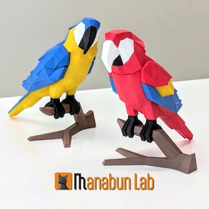 May include: Two geometric parrot sculptures. One is blue and yellow, the other is red, white, and blue. Both are perched on brown branches. The sculptures are on a white surface. The logo "Manabun Lab" is at the bottom.