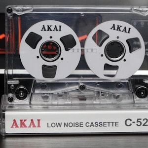 Può includere: Una cassetta audio trasparente in plastica con due bobine nere etichettate "AKAI" e il testo "AKAI LOW NOISE CASSETTE C-52LN" sul fondo.