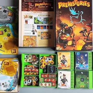 May include: A board game titled "Prehistories" with a box featuring a prehistoric scene. The game includes a game board, cards, and various game pieces. The color palette includes green, brown, yellow, and blue.