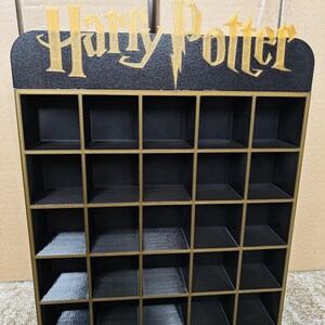 May include: A black and gold display shelf with 25 square compartments. The top features the words "Harry Potter" in gold lettering with a lightning bolt. The shelf is designed to hold small items.