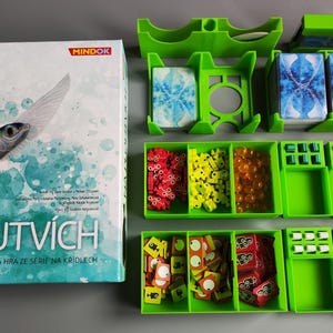 Könnte beinhalten: Brettspielbox mit dem Titel "NA PLOUTVÍCH" und einer Illustration eines fliegenden Fisches. Das Spiel enthält grüne Aufbewahrungsschalen mit Spielsteinen und Karten. Die Spielsteine sind in verschiedenen Farben.
