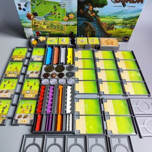 Könnte beinhalten: Eine graue und grüne Brettspielbox mit dem Titel "Naše Osada" und einer Cartoon-Illustration eines Dorfes. Die Box ist geöffnet und die Spielteile sind sichtbar, darunter Holzstücke, Karten und Spielsteine.