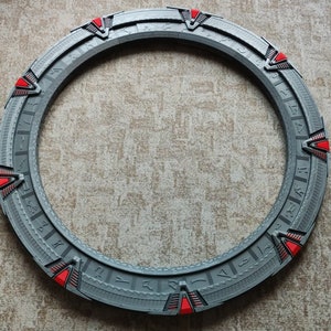 Peut inclure: Un accessoire circulaire gris avec des accents rouges, ressemblant à une porte des étoiles. L'accessoire présente des gravures détaillées et des éléments triangulaires rouges autour du bord extérieur. Le design circulaire est une caractéristique importante.