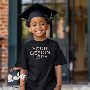 May include: A young person wearing a black graduation cap and gown, smiling and looking at the camera. They are wearing a black t-shirt with the text "YOUR DESIGN HERE" printed on the front.