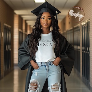 May include: A woman wearing a black graduation gown and cap, a white t-shirt with the text "Add Your Design", and blue jeans. The woman is standing in a hallway with lockers on either side.