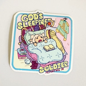 May include: A sticker featuring a cartoon illustration of a sleeping soldier with the text "God's Sleepiest Soldier Reporting In". The soldier is wearing a yellow and white uniform and is lying in bed with a book and a cup of tea. The sticker is on a white background.