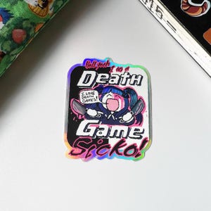 May include: A holographic sticker with a cartoon illustration of a character holding knives, with the text "Hell yeah I'm a Death Game Sicko!" and "I love Death Games!" The sticker has a black and white background.