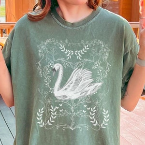 May include: A green oversized t-shirt with a white graphic of a swan in a floral wreath design.