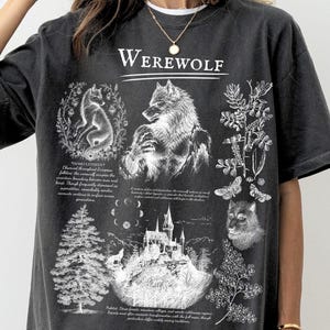 May include: A graphite-colored short-sleeve t-shirt featuring a white graphic print. The design includes the word Werewolf, illustrations of a wolf, a castle, a pine tree, a moth, and lunar phases, accompanied by small blocks of descriptive text.